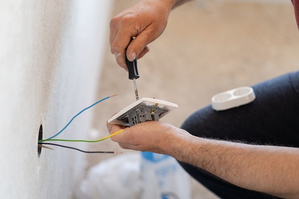 Close up on hands of caucasian man electrician holding screwdriver working on the plug electric on residential electric system installing white AC power socket on gray wall at home repair close up